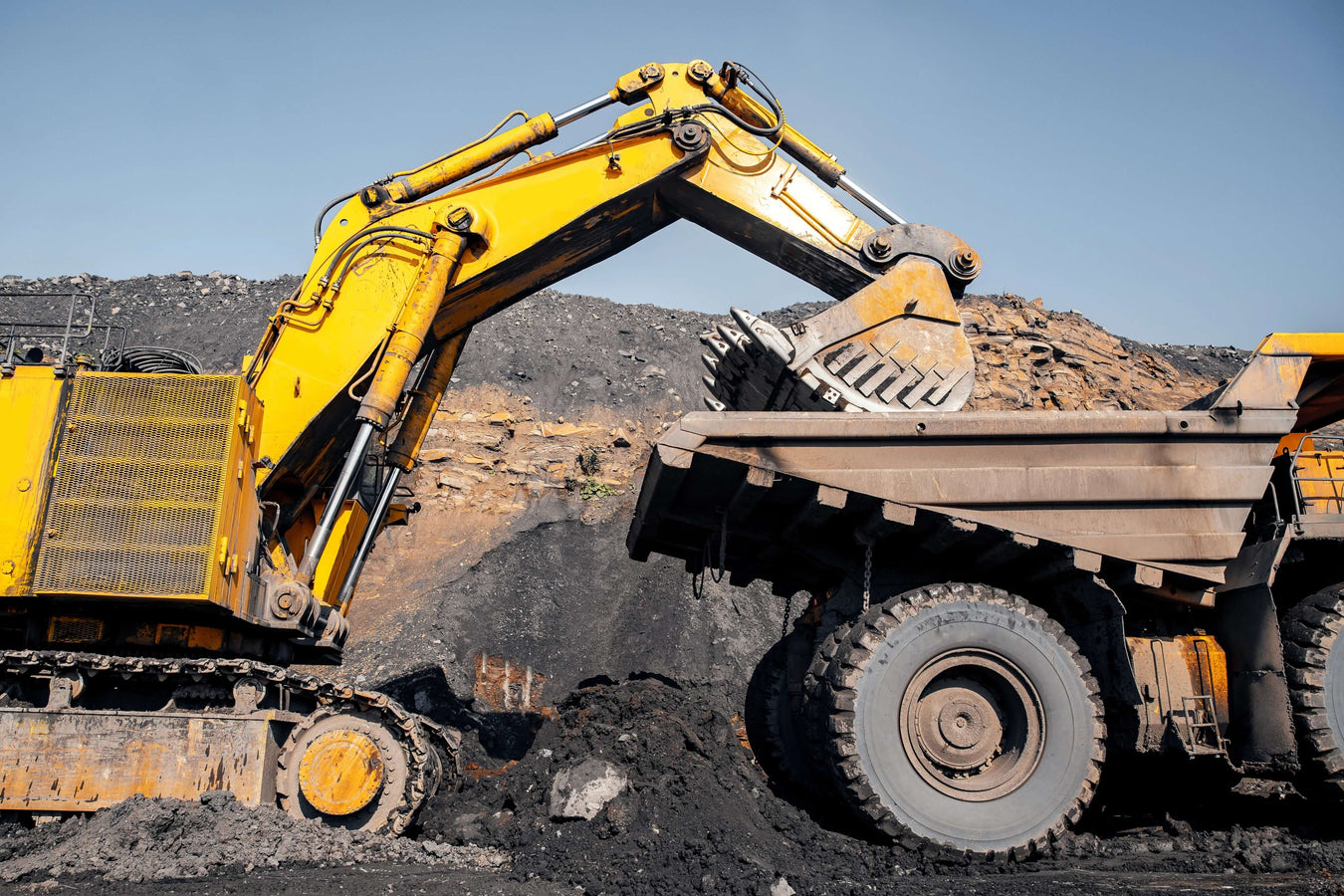 Yellow excavator loading coal into a dump truck against a blue sky.